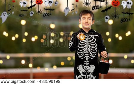 halloween, holiday and trick-or-treating concept - smiling boy in black costume of skeleton with candies and flashlight over roof top party background