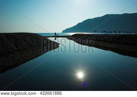 Man is fishing where small river flows into the sea, High contrast silhouette photo against sun, Sun reflecting in water, Parasols on the beach, Adrasan beach on Mediterranean sea in Turkey