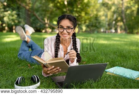 Studentship. Indian Student Girl Learning With Laptop And Book, Taking Notes, Lying In Park Outdoors