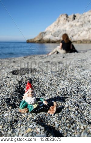 Gnome Lying On The Gravel Of A Beach With A Sitting With Her Back To The Background And The Cliffs O