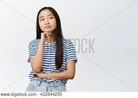 Image Of Asian Girl Thinks, Looks Aside With Thougtful Face Expression, Deciding Smth, Stands Agains