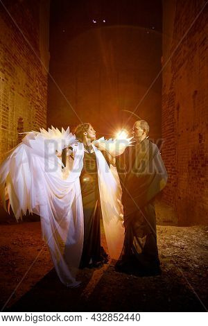 Couple In Dark Clothes In An Abandoned Castle With Old Brick Walls. Man In Black Dress And Woman Wit