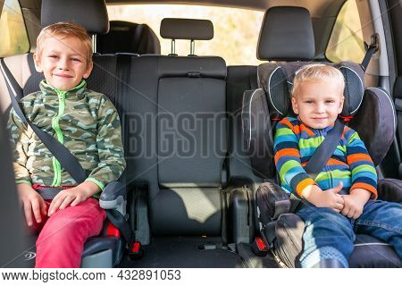 Two Little Boys Sitting On A Car Seat And A Booster Seat Buckled Up In The Car.