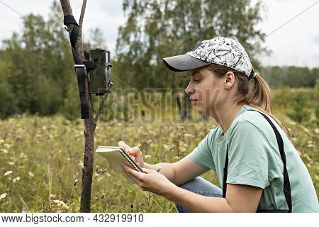 Young Woman Scientist Zoologist Writing Down Data From Trap Camera To Notepad, Observing Wild Animal