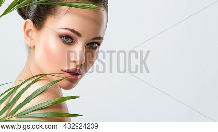 Young beautiful woman with healthy skin of face and palm leaves. Closeup fresh face of an attractive caucasian girl with green plants. Model with bright brown eye makeup. Skin care concept. 