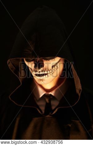 Portrait of the devil, death. Close-up portrait of a dangerous man hiding his face with skull makeup over the hood of a black robe. Black background. Halloween. 