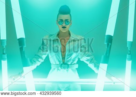 A bright cyberpunk girl in a technological white jumpsuit mirrored sunglasses stands among the neon lights. Portrait of a cool punk rock musician girl in a futuristic cyber style. Studio shot.