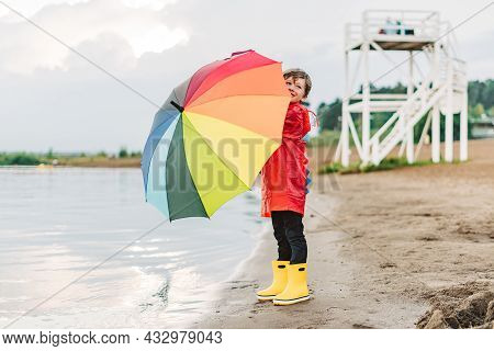 Boy In A Red Raincoat And Yellow Rubber Boots Stands At River Bank And Holding Rainbow Umbrella. Sch