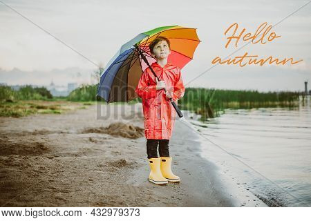 Boy In A Red Raincoat And Yellow Rubber Boots Stands At River Bank And Holding Rainbow Umbrella. Sch