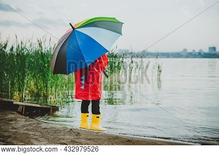 Boy In A Red Raincoat And Yellow Rubber Boots Stands At River Bank And Holding Rainbow Umbrella. Sch