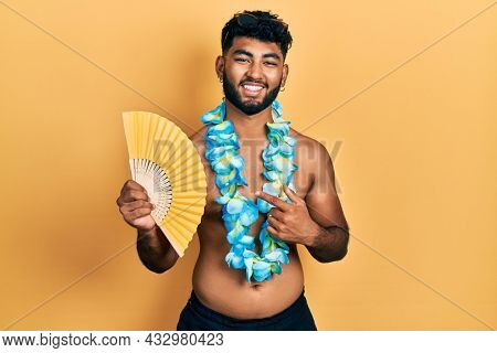 Arab man with beard wearing hawaiian lei holding handfan smiling happy pointing with hand and finger 
