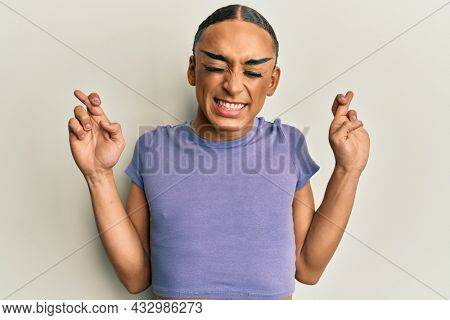 Hispanic man wearing make up and long hair wearing casual t shirt gesturing finger crossed smiling with hope and eyes closed. luck and superstitious concept. 