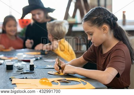 Side view ofo little girl with oil paint preparing halloween paper symbols by table