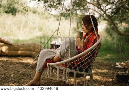 Beautiful Girl Wrapped In Red Plaid Drinking Tea In A Cozy Hanging Chair Outdoors. Girl Holding Mug 