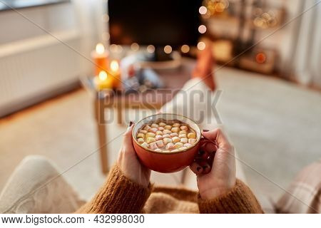 people and leisure concept - young woman watching tv and drinking hot chocolate with marshmallow with her feet on table at cozy home