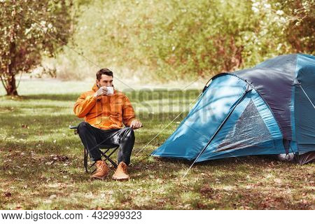 camping, tourism and travel concept - happy man drinking tea at tent camp