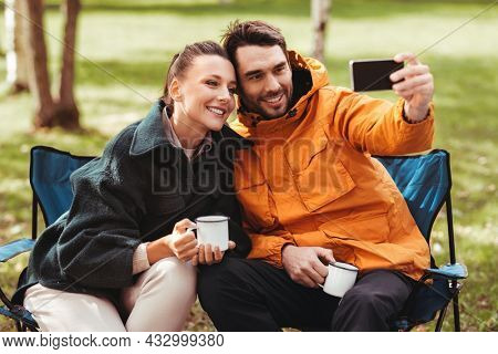camping, tourism and travel concept - happy couple with smartphone drinking tea and taking selfie at tent camp