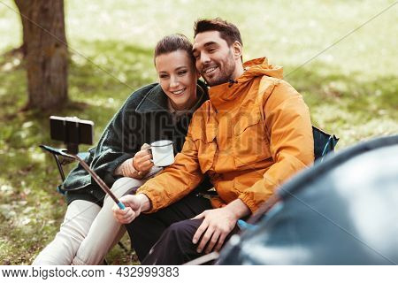 camping, tourism and travel concept - happy couple with smartphone drinking tea and taking selfie at tent camp