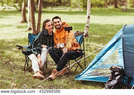 camping, tourism and travel concept - happy couple with smartphone drinking tea and taking selfie at tent camp