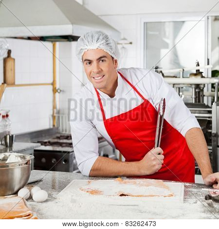 Portrait of smiling male chef holding tongs while preparing ravioli pasta at counter in commercial kitchen