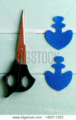 Two elements in the shape of an anchor cut from blue felt. Scissors on wooden background. How to sew a fabric toy for children. Hand crafts. Step. Closeup. Top view