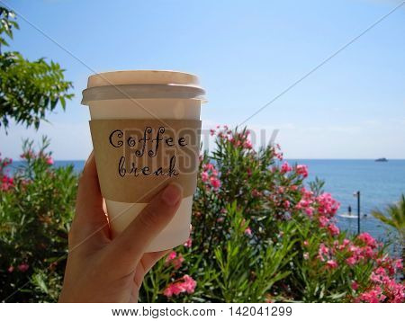 woman's hand holds a coffee Cup that says "break time" in the background of a flowering tree and blue sea