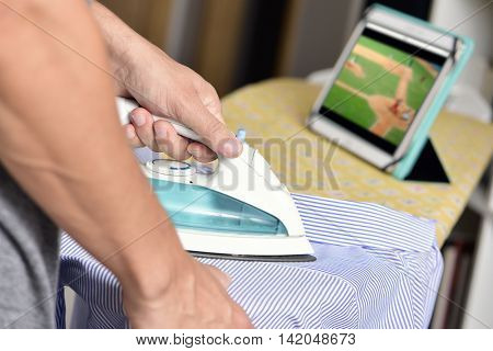 closeup of a young man ironing a striped shirt with an electric iron on an ironing board, while watches a baseball match online in his tablet computer