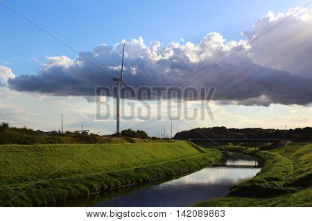 A wind turbine in a peaceful landscape with green grass blue skye and a river