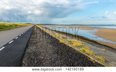 Long asphalt road on an embankment along an estuary in the Netherlands. In the background is a large wind farm. It is early in the morning in the summer season.