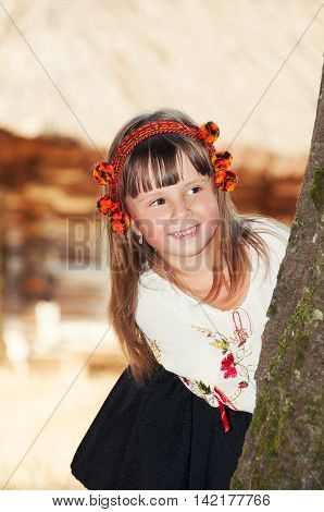 Smiling little girl in the national Ukrainian Hutsul costume and in a wreath looks out from behind a tree
