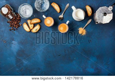 Coffee espresso in cups with italian cantucci, cookies and milk in jug over dark blue painted plywood background, top view, copy space. Food frame concept
