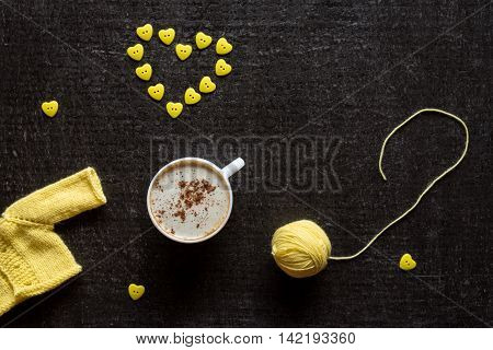 Cup of coffee, yellow yarn and a heart made of buttons on a grunge black background.