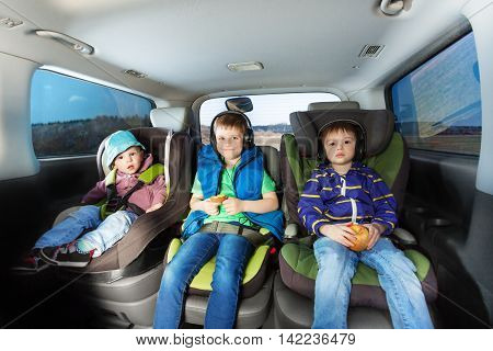 Three happy boys, age-diverse brothers, sitting in safety car seats, snaking and listening the music during car trip
