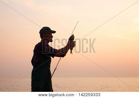 fisherman removes the fish from the hook. old man holding a fishing rod with a caught fish against the setting sun