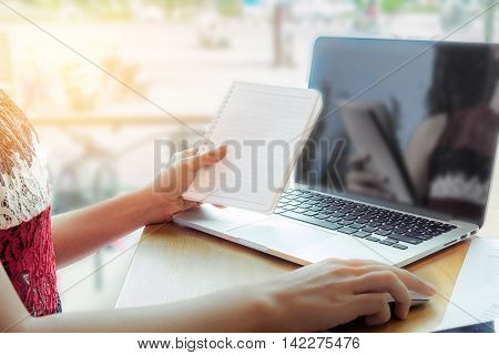 close up hand of young business woman working on laptop computer hand holding notebook and smart phone on wood desk while sitting in coffee shop.soft focus.