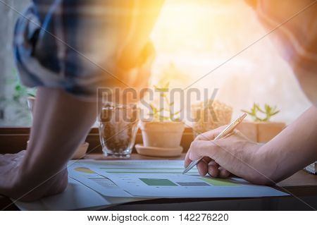 close up hand of business man holding pen and write on paper garph chart on wooden desk indoor office with morning light. vintage filter effect.