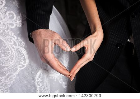 bride's and groom's hands in shape of heart on the background of the bride dress and groom's suit.