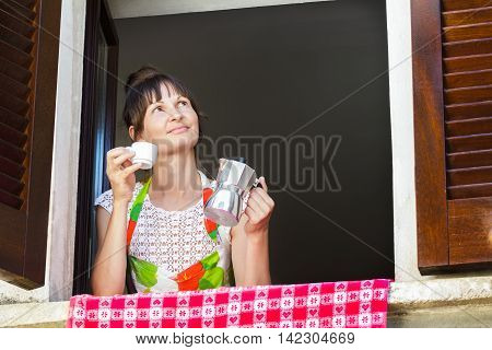 Portrait of middle-aged woman holding white cup of freshly brewed hot coffee and Italian moka pot sitting near open window with traditional European wood brown shutters.