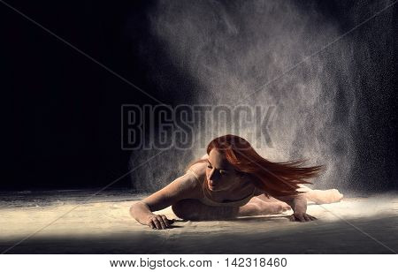 Woman dancer lying on the floor in a ballet pose with flour. Redheaded woman ballerina posing in beige bathing suit on a black background.