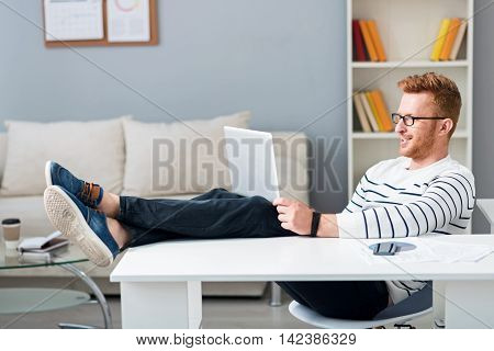 Relaxed atmosphere. Pleasant handsome content man holding his legs on the table and using laptop while working