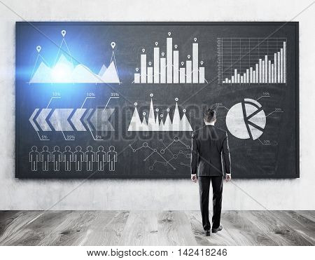 Rear view of man in suit standing in office in front of blackboard with graphs and sketches analyzing data. Concept of stats. Toned image.