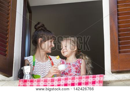 Coffee-break with lollipop sweet of mother with her daughter near open window with traditional European wood brown shutters