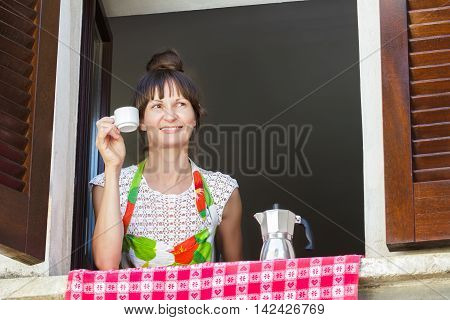 Portrait of smiling woman enjoying little cup of fresh coffee brewed in Italian moka pot sitting near open window with traditional European wood brown shutters.