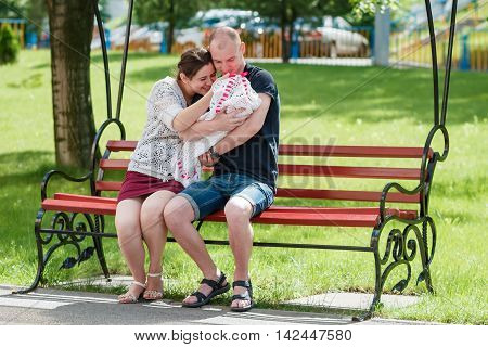 Young Family Sitting On A Park Bench. Father Holding Newborn Daughter In Her Arms.