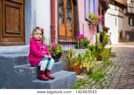 Cute little girl in pink coat walking down a street in historical medieval city center on cold autumn day. Child during fall vacation in Eguisheim Alsace France. Travelling and tourism with kids.