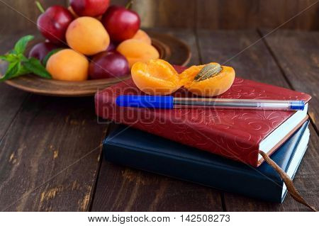 Books for records and a pen in the foregroud. Ripe plums and apricots in a clay bowl in the background. Woode background