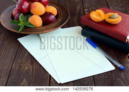 White sheets of paper for your text. Books for records and a pen in the foregroud. Ripe plums and apricots in a clay bowl in the background.
