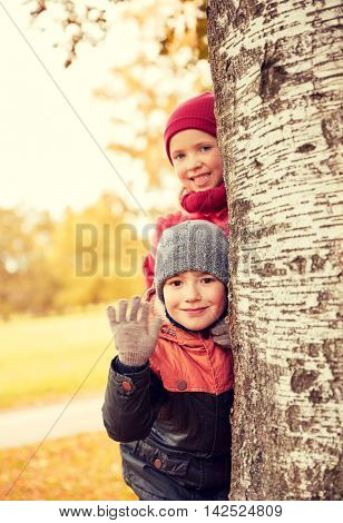 childhood, leisure, friendship, gesture and people concept - happy little girl and boy hiding behind birch tree trunk and waving hand outdoors
