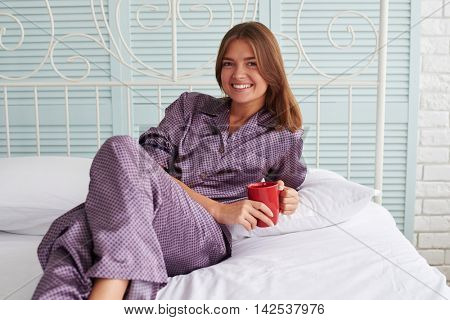Beautiful female is posing at the camera in pajamas holding a cup of coffee and lying on bed in light bedroom with warm domestic atmosphere