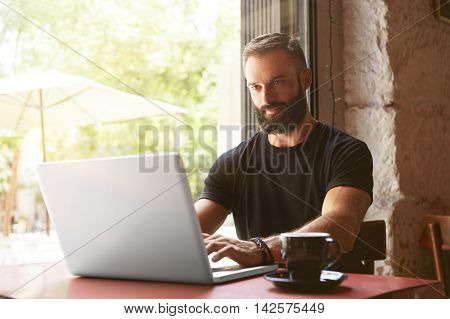 Handsome Bearded Businessman Wearing Black Tshirt Working Laptop Wood Table Urban Cafe.Young Manager Work Notebook Modern Interior Design Place.Coworking Process Business Startup.Blurred Background